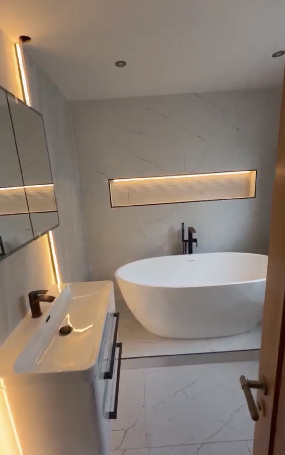 Marble bathroom with a long illuminated niche behind a freestanding bath and lit mirror cabinet above the basin